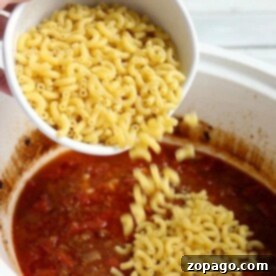 Pouring uncooked elbow macaroni into a simmering pot of crockpot chili mac with a rich tomato base adds hearty texture and delicious flavor.