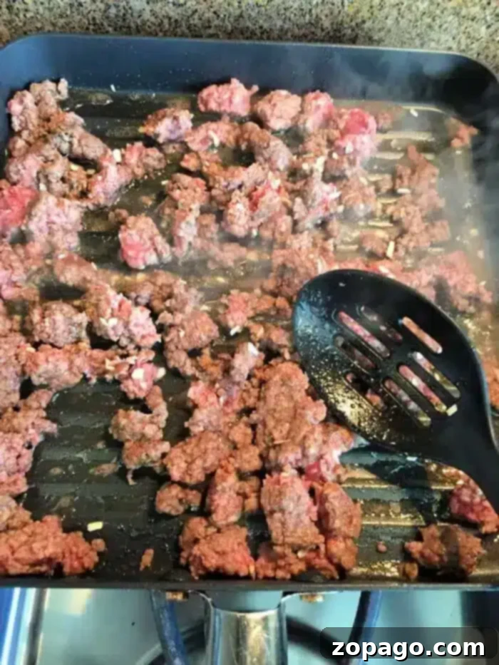 Ground meat sizzles in a grill pan on the stovetop, steam rising as it browns. A slotted black spatula rests atop some of the succulent bits, reminiscent of a 5 ingredient crockpot chili recipe simmering to perfection.