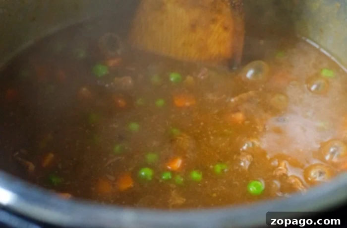 Ultimate Ninja Foodi Stew 5 Stew with peas and carrots simmering in a Ninja Foodi, stirred with a wooden spoon.
