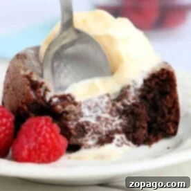 A spoon is cutting into an air fryer chocolate lava cake topped with melting vanilla ice cream on a white plate. Two fresh raspberries sit beside the cake.