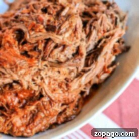 Crockpot BBQ beef, tender and flavorful, sits shredded in a white dish on a table with a red and white striped cloth.