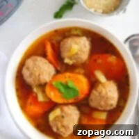 A white bowl contains instant pot meatball soup with sliced carrots and celery, garnished with a parsley leaf. A spoon is on the side, and a small bowl with grated cheese is in the background on a white surface.