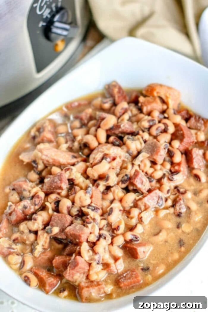 Easy black eyed peas recipe using leftover ham or a ham hock. Homemade tender peas and meat served as a soup or a hearty meal with a side of bread. #crockpotrecipeseasy #crockpotrecipes #blackeyedpeas #blackeyedpeasandham #slowcookerrecipes A rustic wooden bowl of slow-cooker black-eyed peas with ham, served with a spoon, highlighting the texture and richness of the dish.