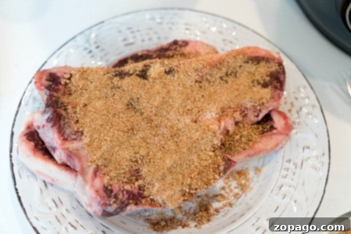 Ultimate Steak Dry Rub 3 A raw steak on a glass plate is generously covered with a dry rub seasoning. The meat features enticing marbling, while the blend of brown spices enhances its appeal. The plate rests elegantly on a light-colored surface.
