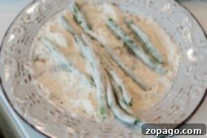 Air Fryer Green Bean Fries coated in flour rest on a decorative white plate, ready to be perfectly crisped by frying or baking.