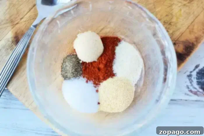 Flavorful Dry Rub Baked Pork Chops 4 A clear glass bowl showcases an array of dry rub spices for perfect baked pork chops: black pepper, paprika, salt, garlic powder, onion powder, and brown sugar. A fork and knife rest nearby on a wooden cutting board.