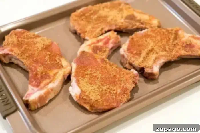 Flavorful Dry Rub Baked Pork Chops 7 Four exquisitely seasoned pork chops, promising to be the best baked pork chops you've ever tasted, are arranged on a baking sheet, liberally sprinkled with a rich brown spice blend, ready for baking on a light-colored surface.