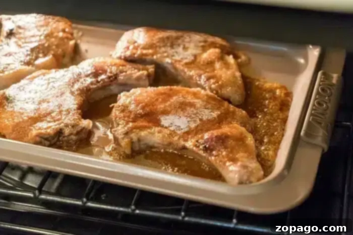 Flavorful Dry Rub Baked Pork Chops 8 Perfectly baked pork chops sit on a metal baking tray inside an oven, glistening with golden-brown seasoning and natural juices. The tray handle, with the "Anolon" brand visible, completes the image of the best baked pork chops.