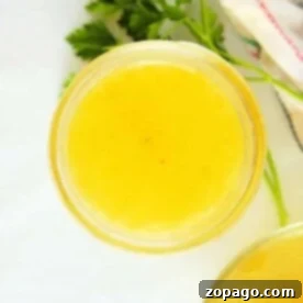 Top view of a glass jar filled with yellow liquid, resembling chicken broth, on a white background with a parsley sprig nearby.