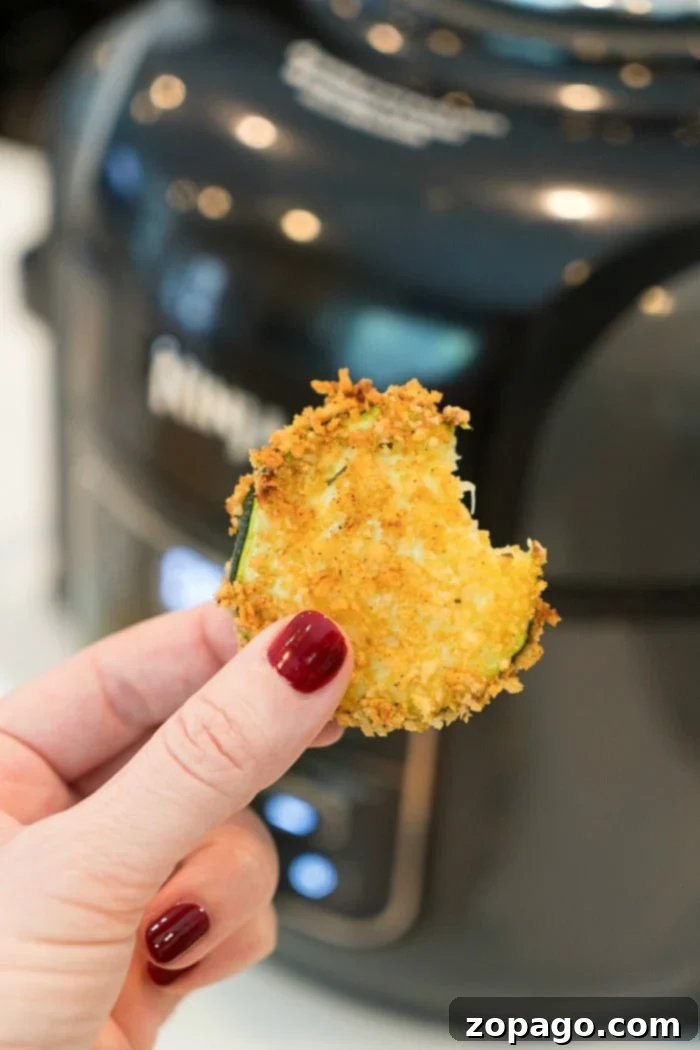 Crispy Low-Carb Air Fryer Meals 8 A hand holding a perfectly crispy, lightly breaded zucchini chip in front of an air fryer, with a plate of freshly made air fryer zucchini chips visible, highlighting a delicious and healthy snack option.