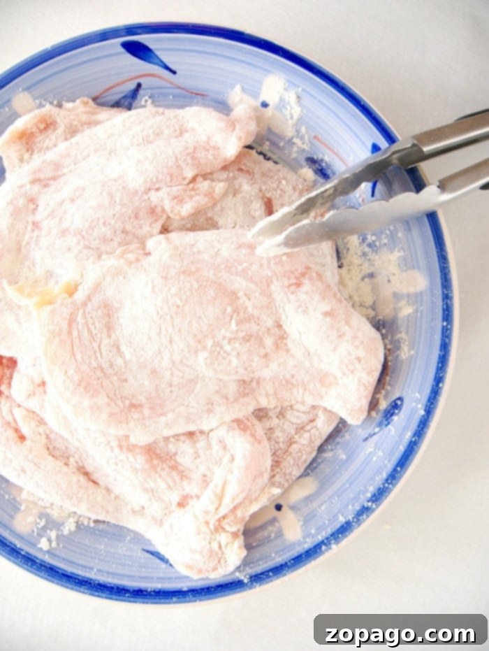 Raw chicken cutlet pieces, lightly coated in flour, ready for cooking on a blue plate with metal tongs.