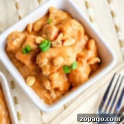 White bowl filled with creamy peanut butter chicken, topped with peanuts and green onions. Fork and napkin beside the bowl, ready to enjoy this delicious peanut butter chicken recipe.