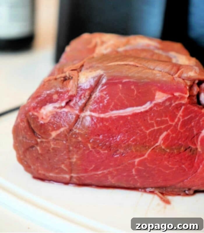 Crispy Air Fryer Roasts 2 A close-up image of a raw beef roast destined for the air fryer, resting on a white surface. The marbled texture and vibrant red color of the meat are prominently displayed against a dark, blurred background.