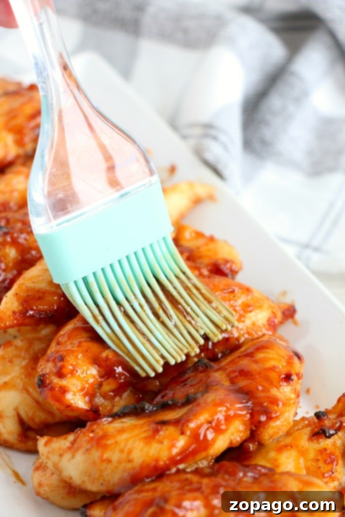 Tender and juicy air fryer chicken tenders without breading, being brushed with extra barbecue sauce. A brush applies barbecue sauce to freshly cooked air fryer chicken tenders on a white plate, highlighting a simple, no breading meal.