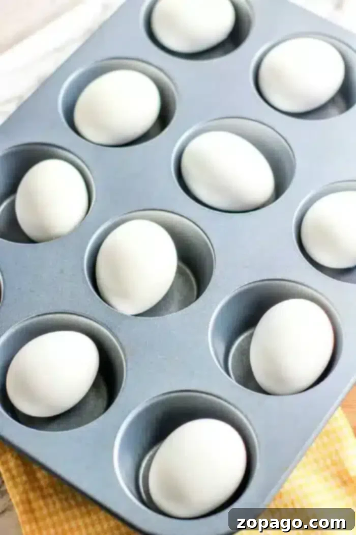 Baked Hard Boiled Eggs 2 A muffin pan with nine white eggs placed in the cups sits atop a yellow checkered cloth, demonstrating how to hard boil eggs in the oven with ease.