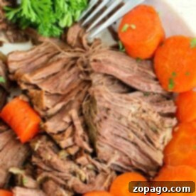 Perfectly Tender Instant Pot Frozen Roast 7 Close-up of a plate with shredded cooked beef, perfectly tender from being a frozen roast in the Instant Pot, surrounded by sliced, cooked carrots and a sprig of fresh parsley. A fork is placed beside the beef, ready to serve.