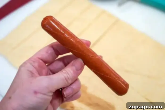 Perfectly Crispy Air Fryer Pigs in a Blanket 6 A hand holds a raw hot dog, already scored, and a strip of crescent dough ready to be wrapped for air frying.