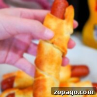 Perfectly Crispy Air Fryer Pigs in a Blanket 9 A hand holds a baked crescent roll wrapped around a hot dog, reminiscent of classic air fryer pigs in a blanket. In the background, more wrapped hot dogs rest on a white plate. A bottle of mustard is partially visible to the right.