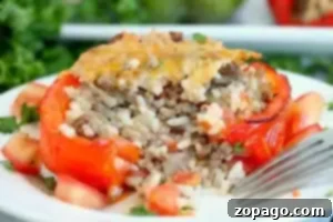 A stuffed red bell pepper, blanched to perfection, sits on a white plate filled with a savory mix of ground meat and rice, topped with melted cheese. Diced tomatoes and chopped herbs are artfully scattered around the pepper, while a fork rests beside it.