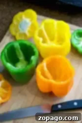 A wooden cutting board displays hollowed-out bell peppers in vibrant yellow, green, and orange hues. A sharp knife with a black handle lies beside them, perfect for prepping ingredients in your kitchen adventures, whether slicing or learning how to blanch bell peppers.