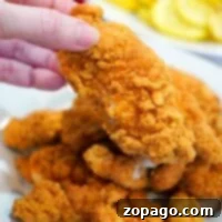 A hand holding a piece of crispy fried chicken above a plate of golden brown chicken pieces cooked to perfection, just like frozen chicken tenders prepared in an air fryer.
