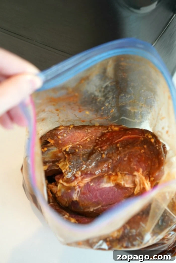 Cook Once, Eat All Week 8 A hand holds an open plastic bag containing raw meat marinated in a red sauce with visible garlic pieces, ready for the air fryer, on a light-colored surface.