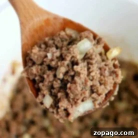 A close-up of crockpot ground beef with onions rests on a wooden spoon above a white bowl, showcasing its rich texture and savory aroma.