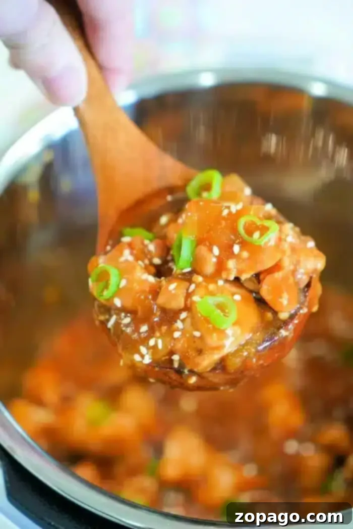 Easy Instant Pot Korean Chicken 2 A close-up of a wooden spoon holding a serving of an Instant Pot Korean chicken dish with diced vegetables in a thick sauce. The dish is garnished with sesame seeds and sliced green onions. The background shows a pot containing the rest of the flavorful meal.