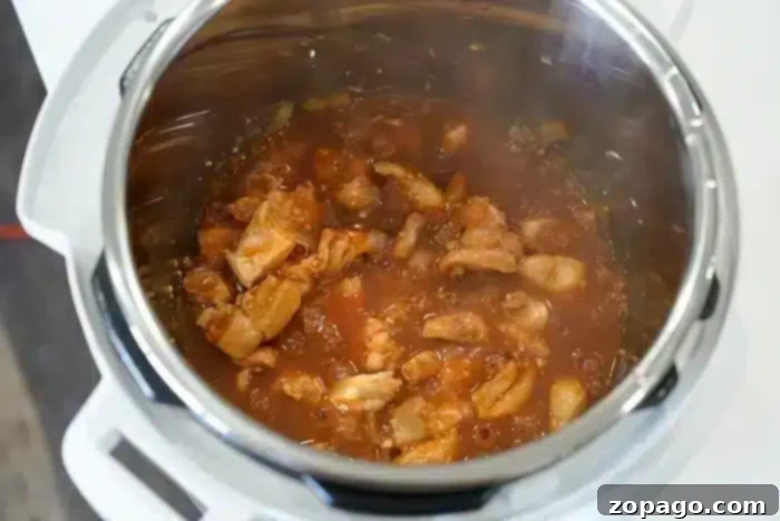 Easy Instant Pot Korean Chicken 6 A close-up view of an Instant Pot containing chicken pieces simmering in a red tomato-based sauce. The dish, possibly Korean chicken, appears to be in the process of cooking, with visible steam and some chopped onions mixed in.