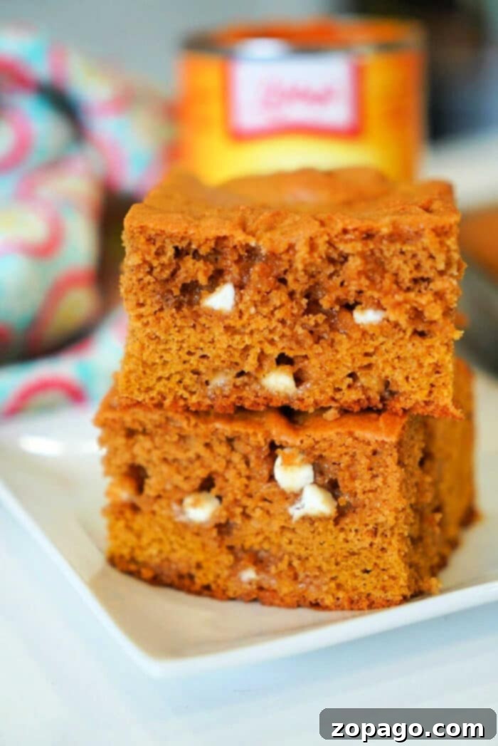 Golden Harvest Pumpkin Blondies 2 Two delicious pumpkin blondies with white chocolate chips are stacked invitingly on a pristine white plate, set against a vibrant, colorful cloth in the background.