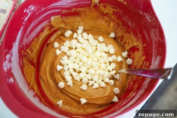 Golden Harvest Pumpkin Bars 8 A red mixing bowl brimming with dough and white chocolate chips promises delightful pumpkin blondies, while a metal spoon rests inside, ready for action.