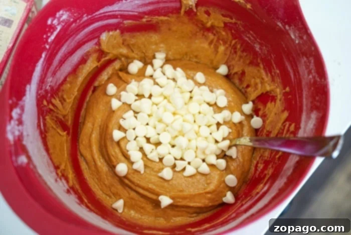 Golden Harvest Pumpkin Blondies 9 A vibrant red mixing bowl filled with pumpkin blondie batter, generously studded with white chocolate chips, with a metal spoon resting inside, ready for the next step.