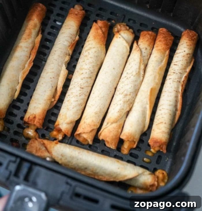 Crispy Air Fryer Frozen Taquitos 2 An air fryer basket contains several frozen taquitos arranged in neat rows, now transformed into crispy delights. Each taquito is golden brown with some browned edges, and a bit of cheese has melted out from the ends.