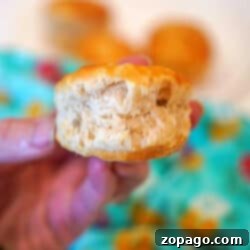 A hand holding a fluffy biscuit with a bite taken out, fresh from frozen biscuits in air fryer, with more biscuits and a turquoise cloth in the background.