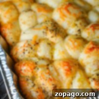 Smoky Traeger Pull-Apart Loaf 12 A close-up of a baked dish in a foil tray, showcasing golden-brown smoked bread covered with melted cheese and sprinkled with herbs. The bread appears fluffy and slightly crispy on top, with unevenly distributed cheese and herbs.