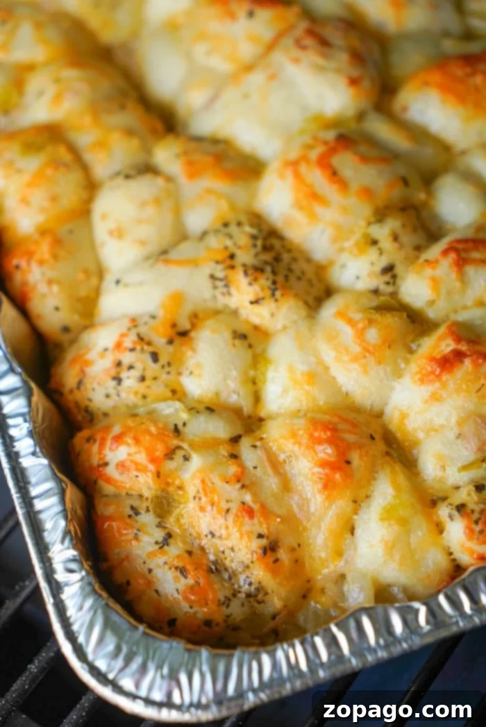 Smoky Traeger Pull-Apart Loaf 14 A close-up of a baked dish in a foil tray, showcasing golden-brown smoked bread covered with melted cheese and sprinkled with herbs. The bread appears fluffy and slightly crispy on top, with unevenly distributed cheese and herbs, ready to be pulled apart.