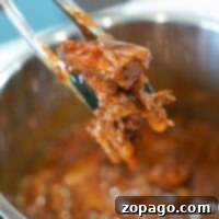 Tongs holding flavorful, shredded Instant Pot BBQ beef above a pot of simmering sauce.