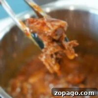 Tongs holding flavorful, shredded Instant Pot BBQ beef above a pot of simmering sauce.
