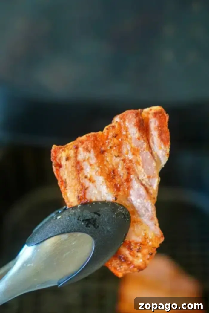 Crispy Air Fryer Pork Belly 2 A piece of seasoned and cooked air fryer pork belly is being held by a pair of metal tongs. The background is slightly blurred and appears to be an air fryer. The pork belly is visibly crispy and browned around the edges.