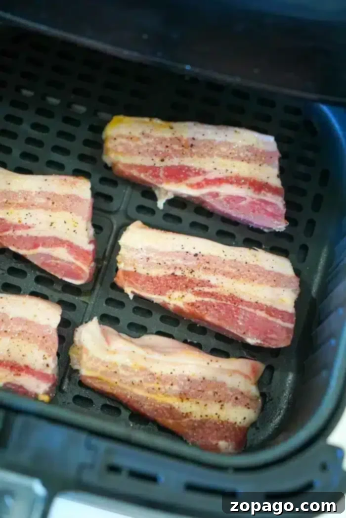 Crispy Air Fryer Pork Belly 5 Six strips of raw bacon are arranged on the tray inside an air fryer, ready to be cooked. The bacon strips, seasoned with black pepper and salt, resemble thin pieces of pork belly. The air fryer tray has a grid pattern for even cooking.