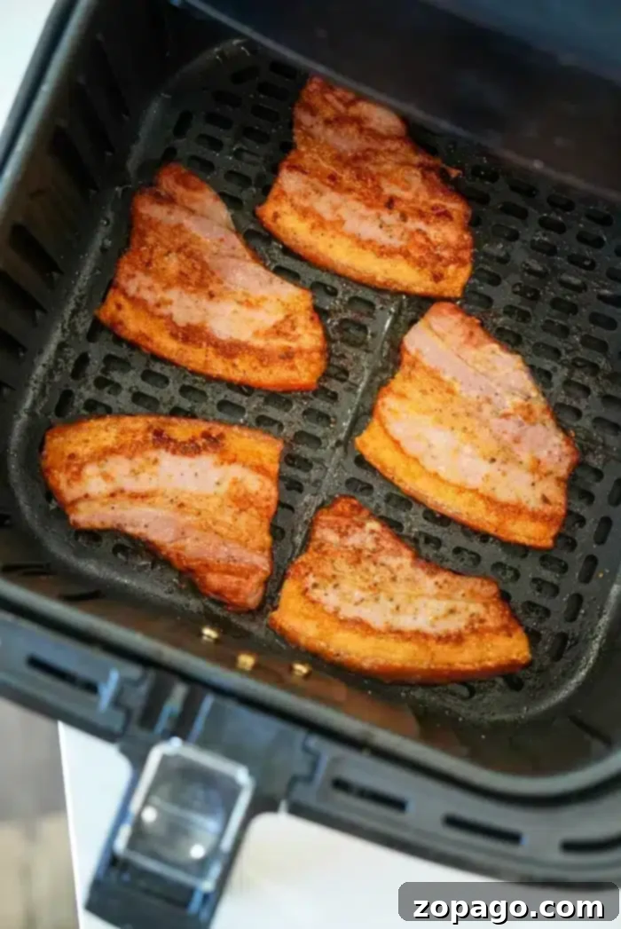 Crispy Air Fryer Pork Belly 7 Six pieces of pork belly bacon, partially cooked with browned edges, are arranged inside an air fryer basket. The basket has a black grid-like base and sits on a white surface.