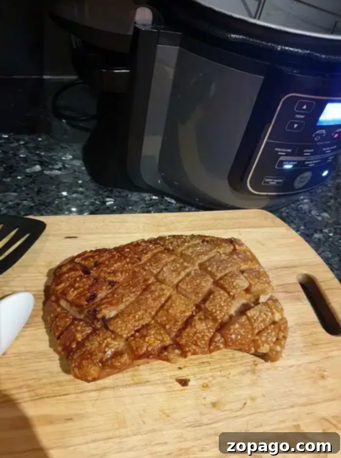 Crispy Air Fryer Pork Belly 8 A wooden cutting board holds a piece of cooked pork belly with crispy, cross-hatched crackling. A spatula handle is visible on the left, and a slow cooker with digital controls sits in the background on a dark kitchen countertop, highlighting the results of perfectly air-fried pork belly.