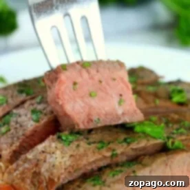 A fork holds a slice of medium-rare steak garnished with chopped herbs. Several slices, cooked from frozen in an air fryer, are arranged on a plate in the background, also sprinkled with herbs, against a backdrop of blurred green foliage.