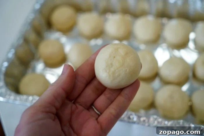 A person delicately holds a small, round dough ball above a tray of similar dough balls, each destined to become a piece of aromatic smoked bread.