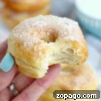 A hand holds a bitten, sugar-dusted cronut, reminiscent of a delightful fusion between biscuit and donut, with more cronuts stacked enticingly in the background.