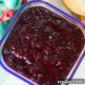 A square container filled with rich huckleberry jam sits next to a piece of bread and a colorful floral cloth.