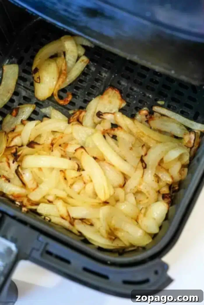 Golden Crisp Air Fryer Onions 2 Sliced onions are shown cooking in an air fryer basket, partially browned and in the process of caramelizing. The air fryer is open to display the air fryer onions inside.