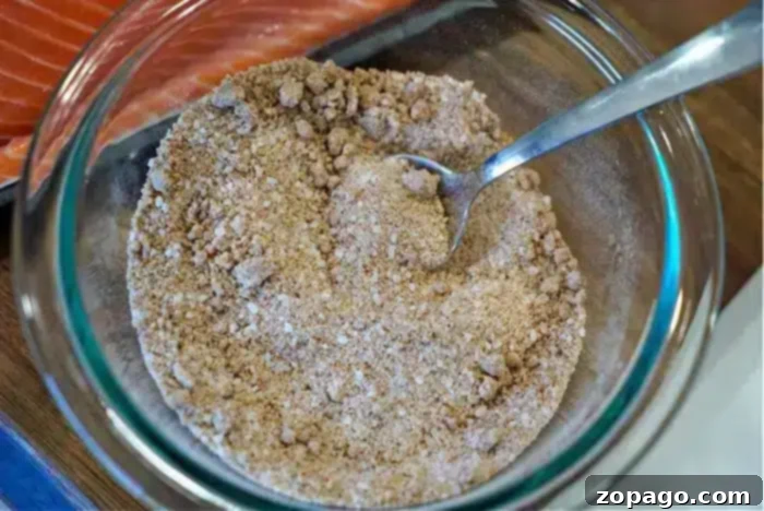 Ultimate Baked Salmon 4 A glass bowl holds a textured, brown dry seasoning mix with a spoon nestled inside. In the background, a portion of raw salmon sits ready for transformation into the best baked salmon.