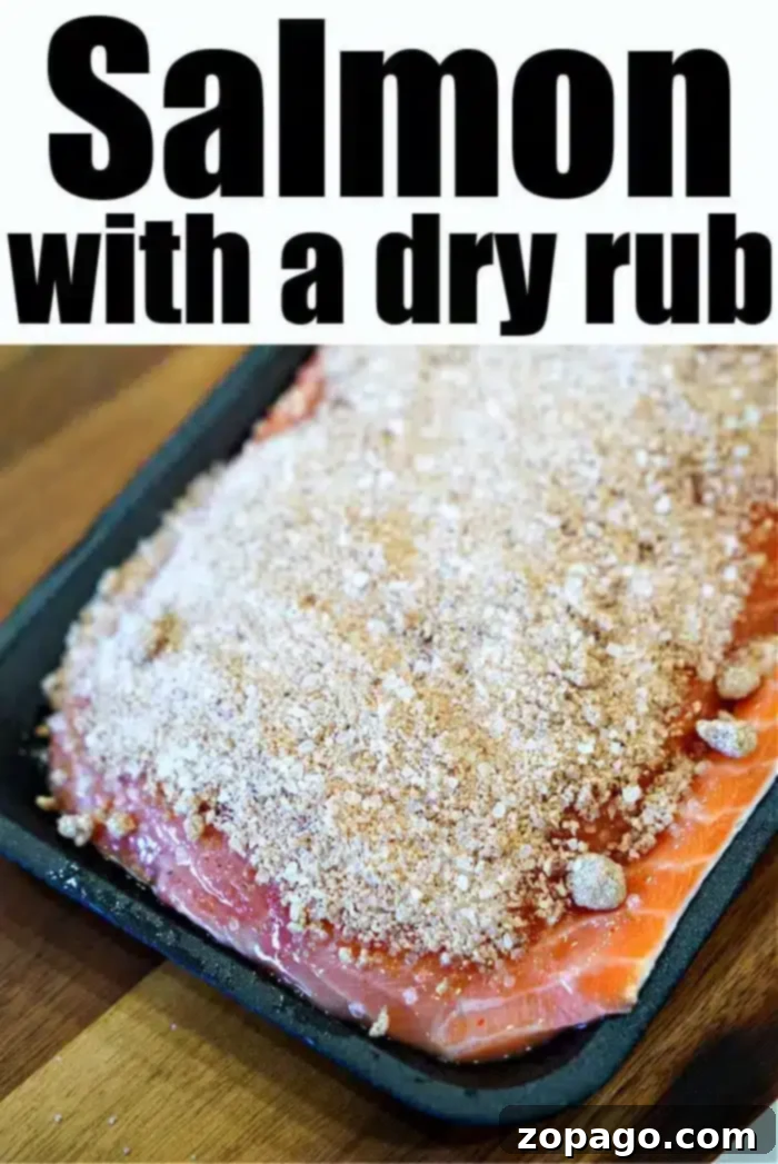 Ultimate Baked Salmon 6 A raw salmon fillet, touted as the best baked salmon, is covered with a dry rub seasoning on a black tray. The image displays "Salmon with a dry rub" at the top in bold letters. The tray rests on a wooden surface.