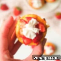 A hand holds a mini strawberry cheesecake topped with cream, perfectly sized from being baked in muffin tins. In the background, more cheesecakes rest invitingly on a plate.